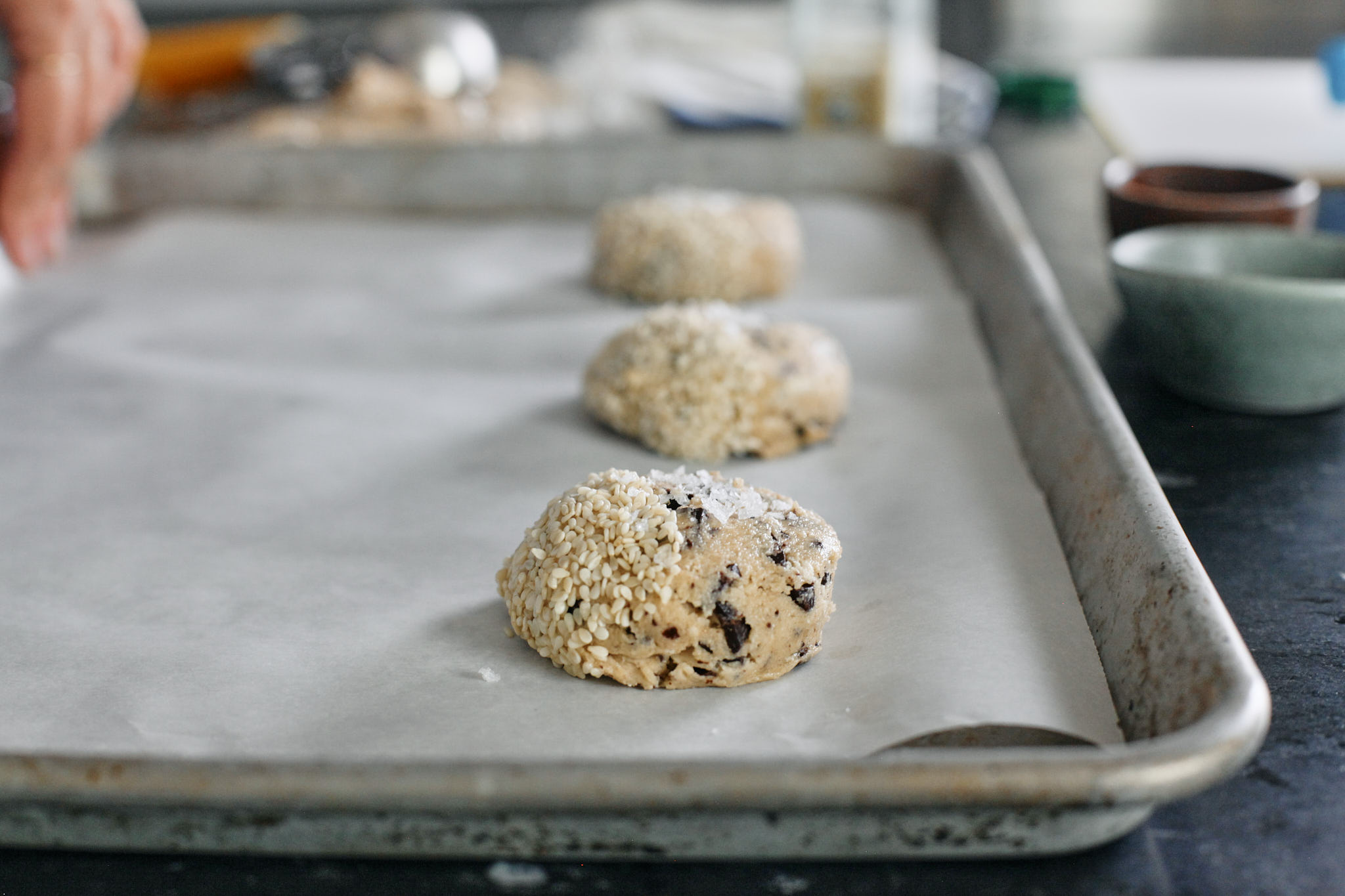 halva chocolate chip cookie dough scooped and placed on a a parchment lined cookie sheet, half topped with sesame seeds and half topped with flaky sea salt