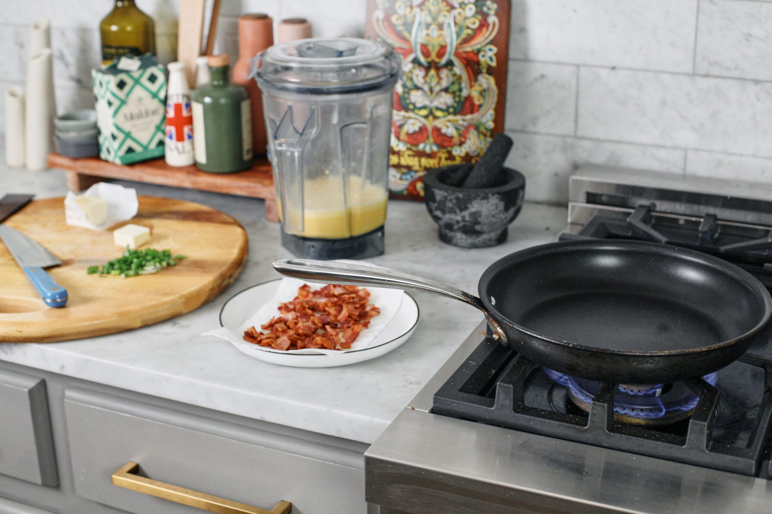 Bacon and Egg Sheet Pan Tacos 1 Kitchen counter and stovetop. There's a cutting board, blender, and plate of cooked bacon on the countertop, and an empty skillet on the stove.