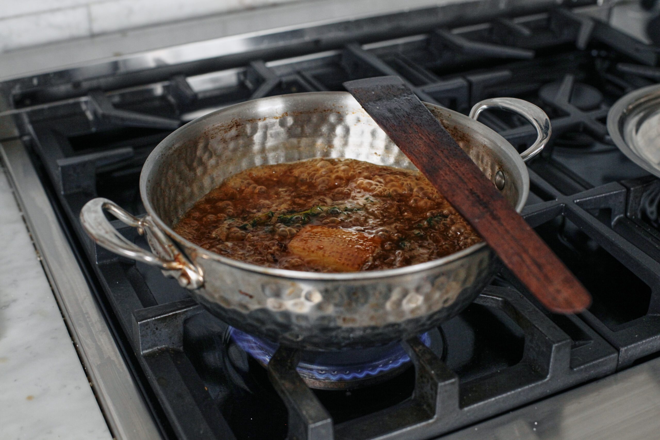 broth simmering with a parmesan rind and thyme in a hammered steel sauté pan on a gas range with a wooden spatula resting on the pan