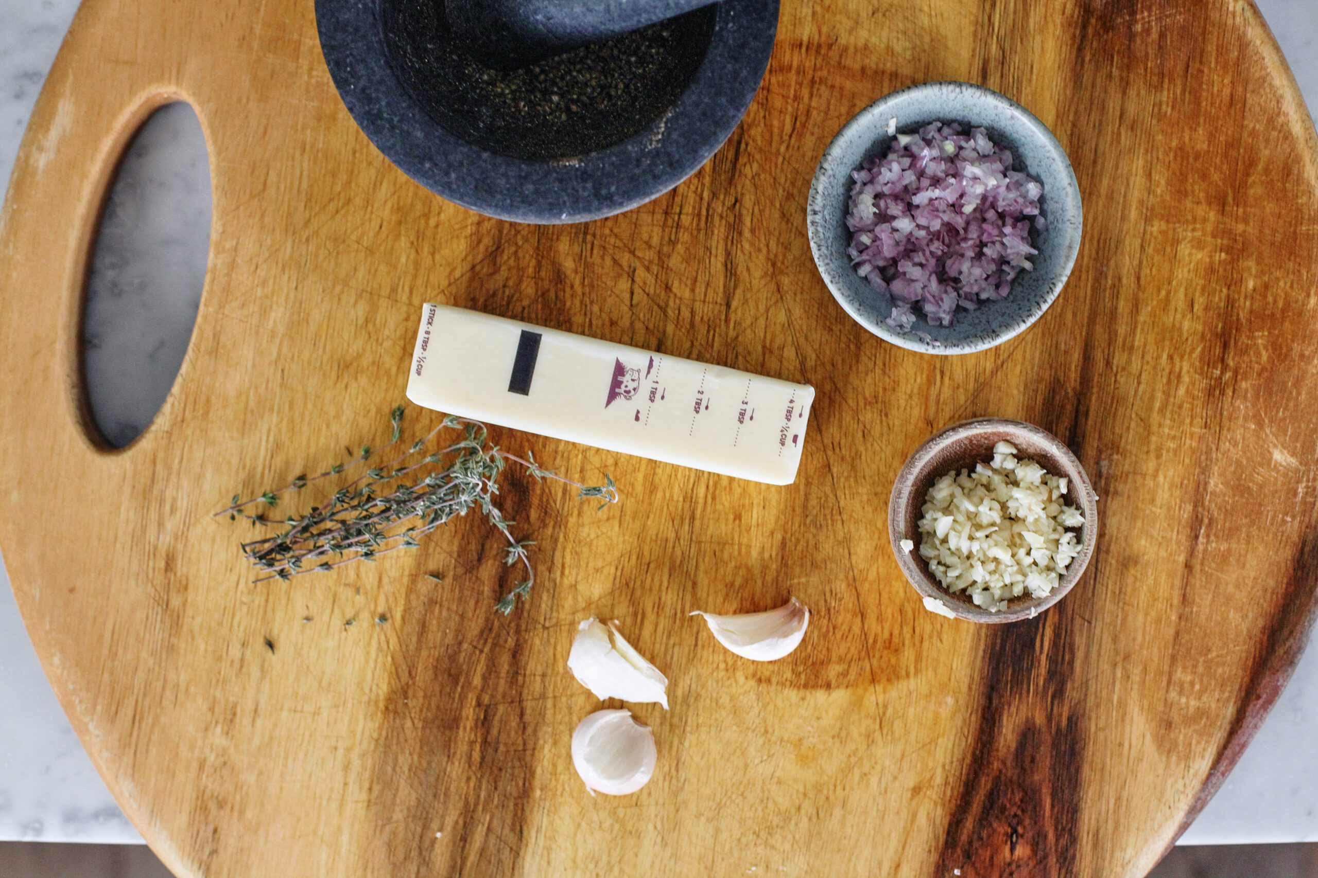 overhead photo of a round wooden cutting board with a mortar and pestle that has black peppercorns in it, a stick of butter, small bowls of minced shallot and garlic, sprigs of fresh thyme, and three unpeeled garlic cloves