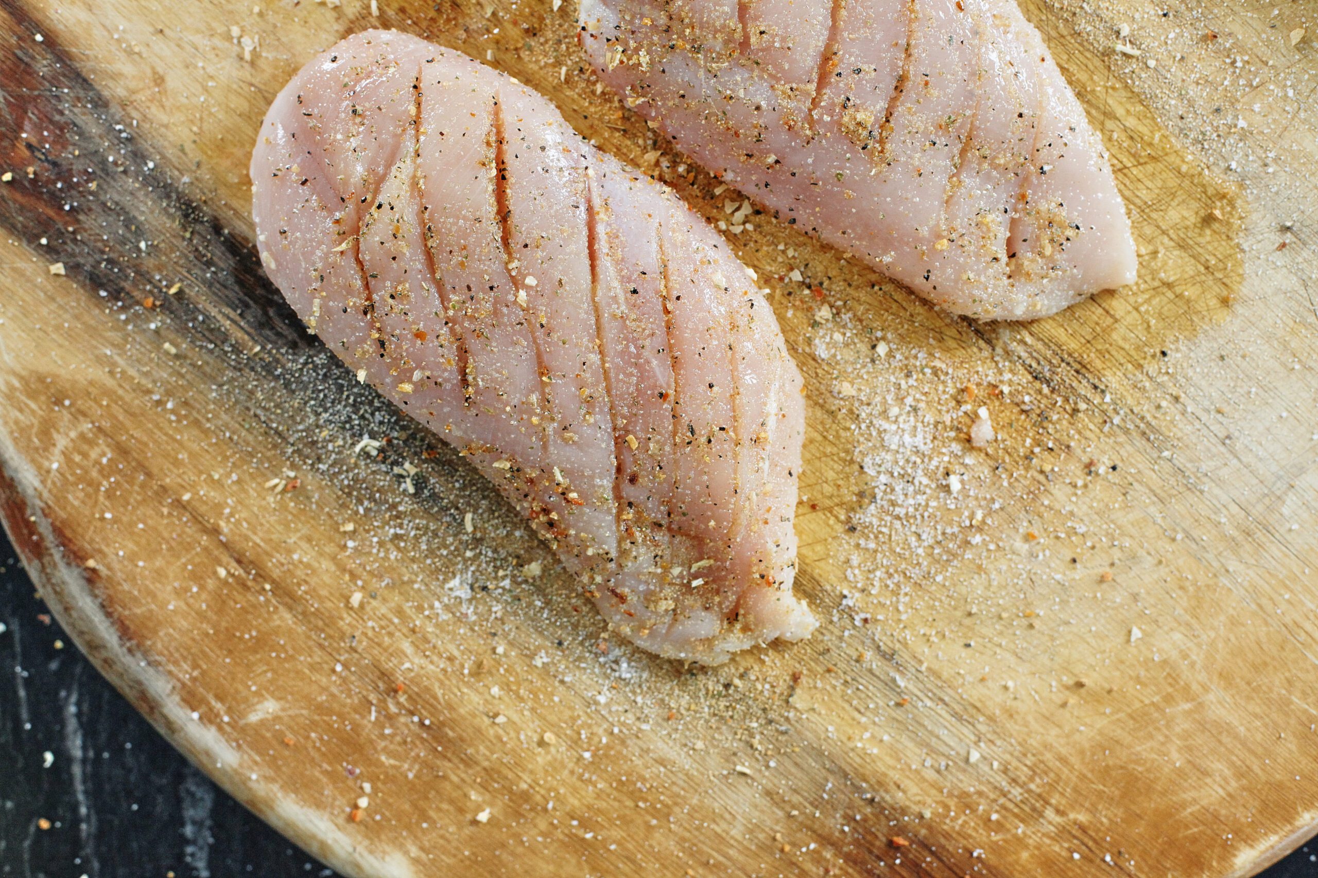 two raw chicken breasts on a wooden cutting board with seasonings on them