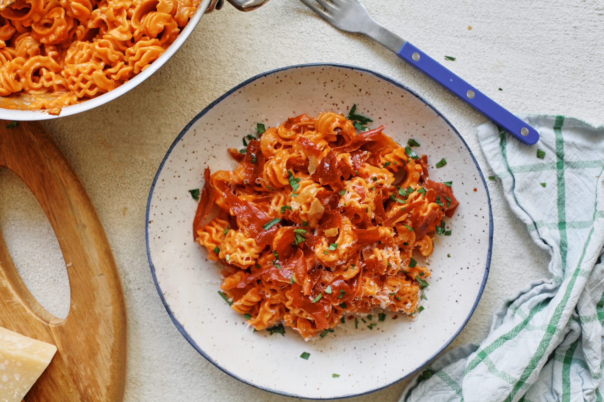 overhead photo of spicy vodka radiatore pasta with pieces crispy prosciutto and Italian parsley on top. It is in a white dish with a black rim, and theres a blue-handled silver fork next to the dish