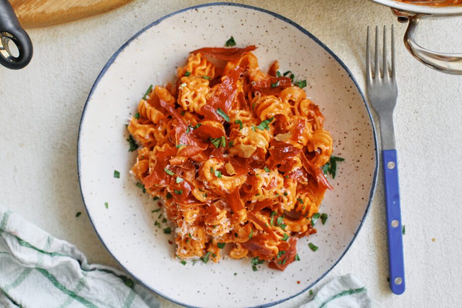 overhead photo of spicy vodka radiatore pasta with pieces crispy prosciutto and Italian parsley on top. It is in a white dish with a black rim, and theres a blue-handled silver fork next to the dish