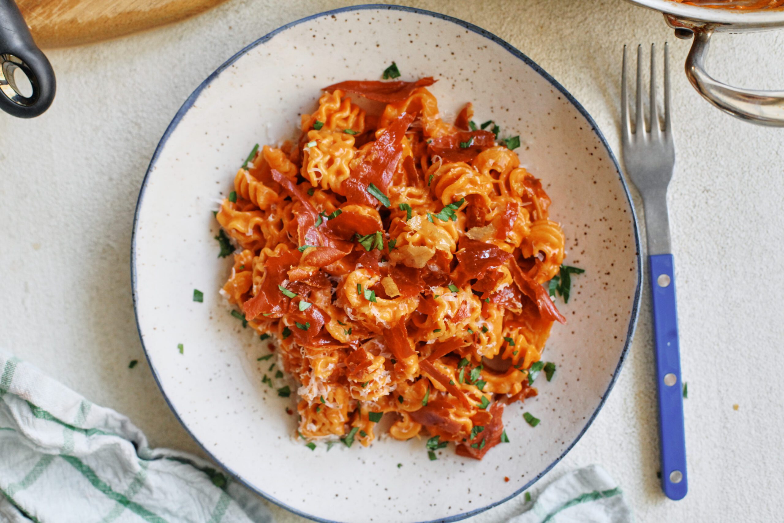 overhead photo of spicy vodka radiatore pasta with pieces crispy prosciutto and Italian parsley on top. It is in a white dish with a black rim, and theres a blue-handled silver fork next to the dish