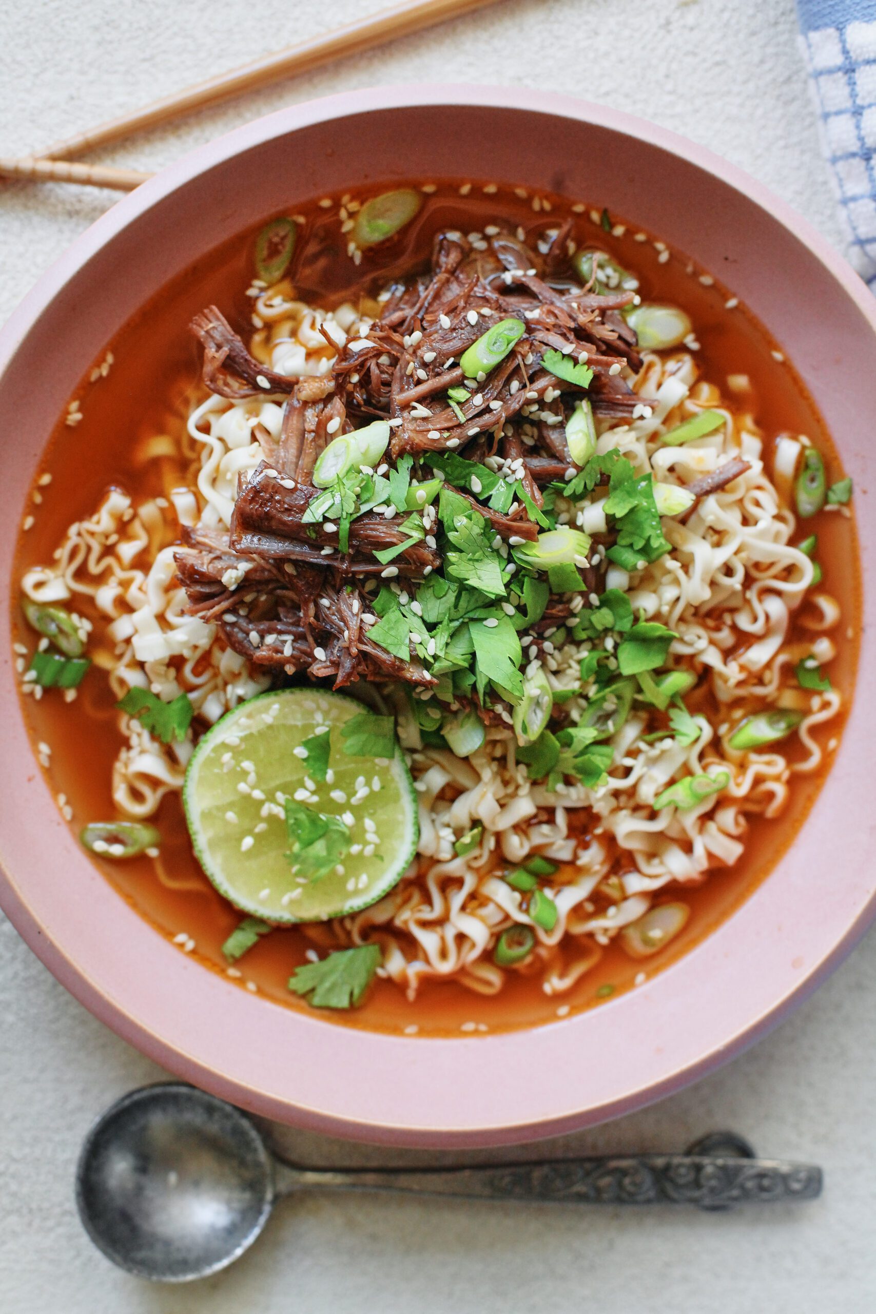 beef short rib noodle soup in a light pink bowl, garnished with half a lime, sesame seeds, and cilantro