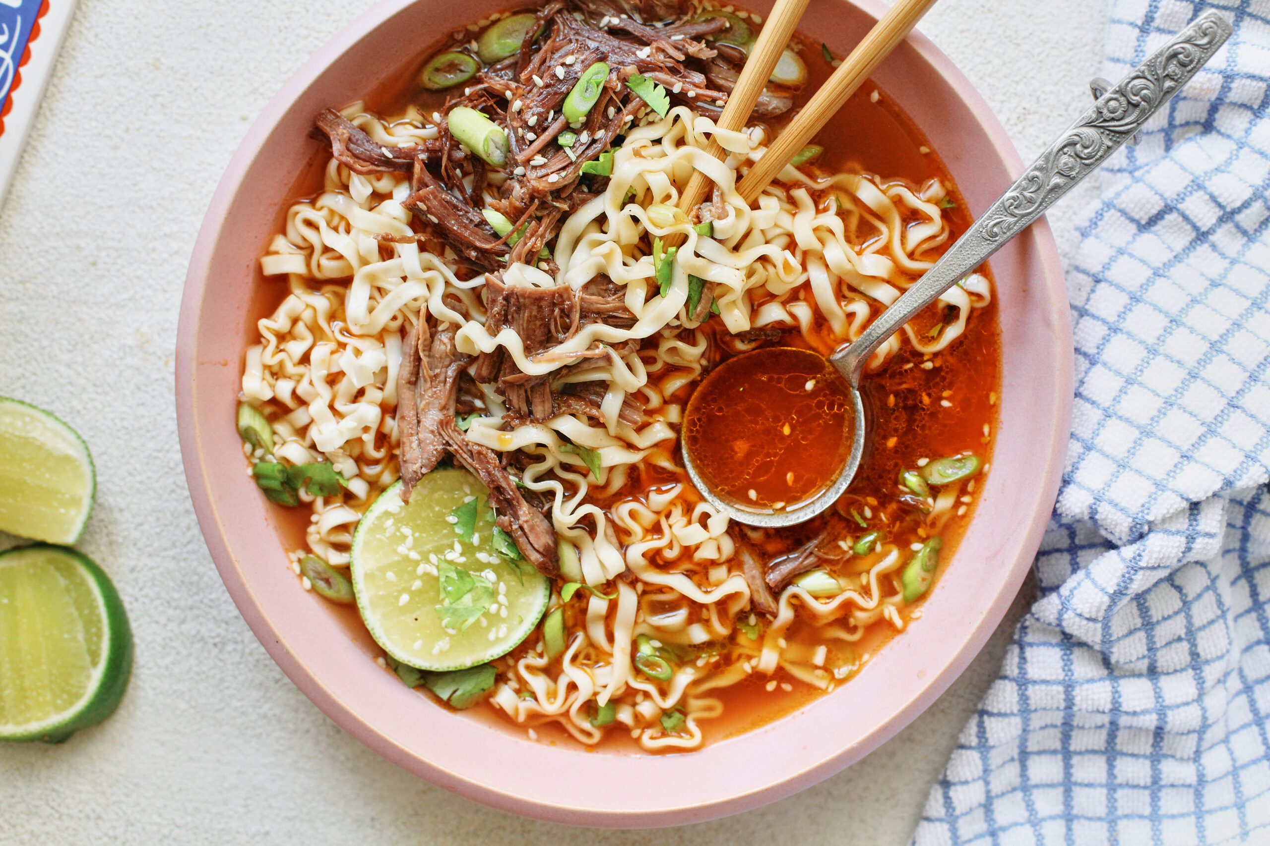 beef short rib noodle soup in a light pink bowl, garnished with half a lime, sesame seeds, and cilantro. there are wooden chopsticks and a silver spoon in the bowl. the spoon is filled with red-orange broth