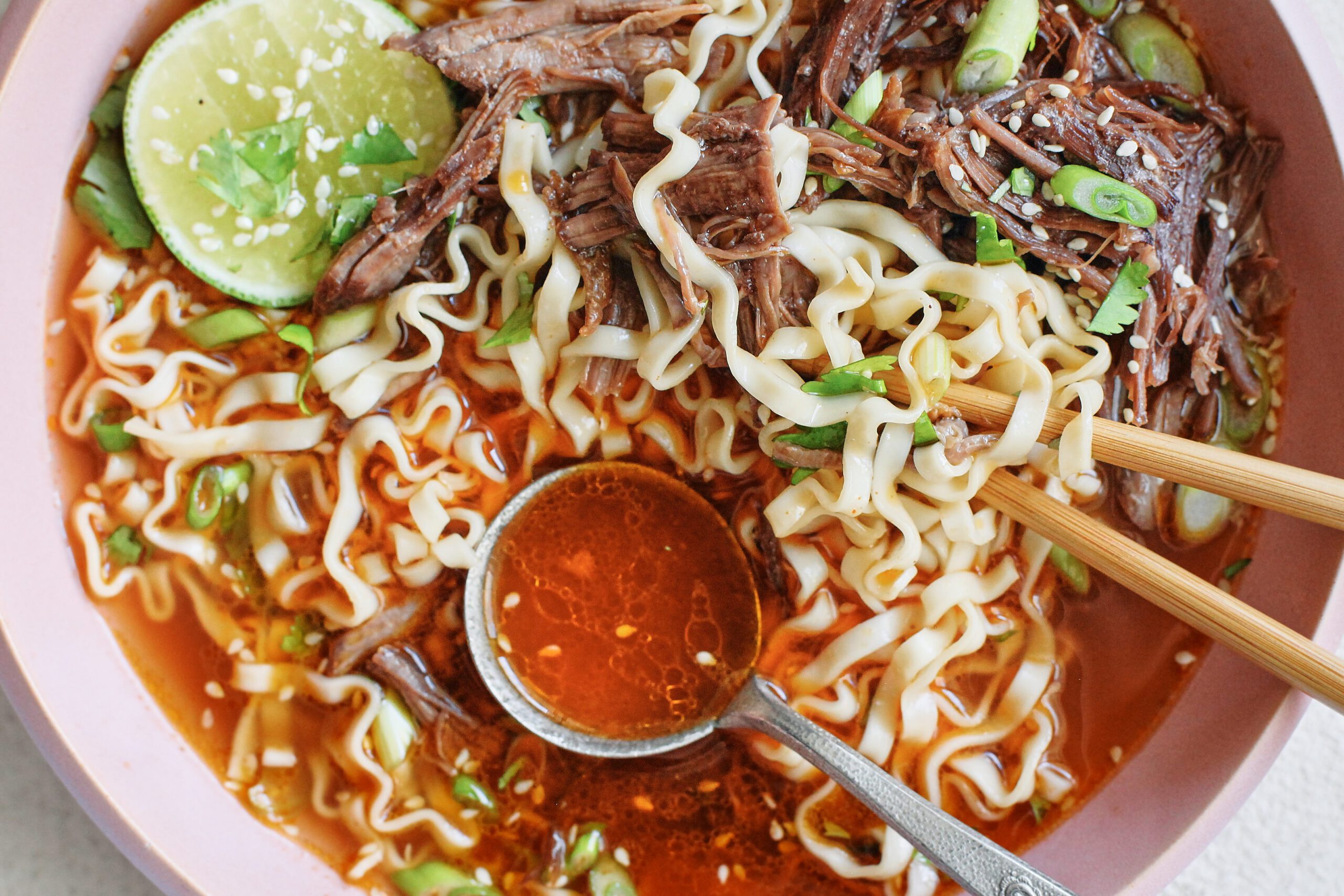 beef short rib noodle soup in a light pink bowl, garnished with half a lime, sesame seeds, and cilantro. there are wooden chopsticks and a silver spoon in the bowl. the spoon is filled with red-orange broth