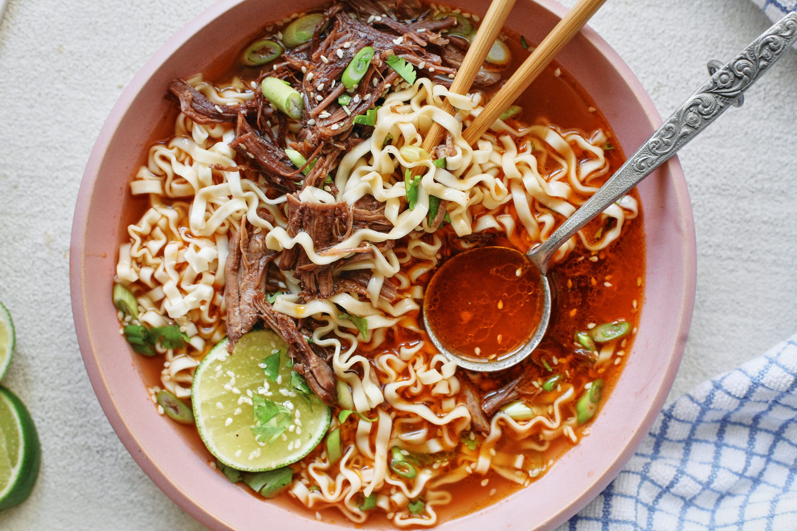 beef short rib noodle soup in a light pink bowl, garnished with half a lime, sesame seeds, and cilantro. there are wooden chopsticks and a silver spoon in the bowl. the spoon is filled with red-orange broth