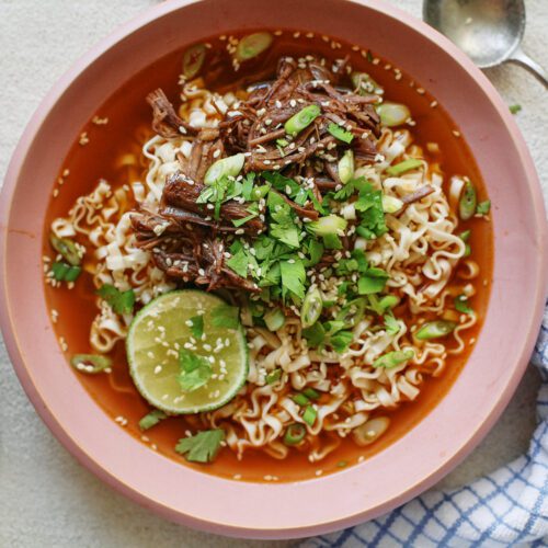 beef short rib noodle soup in a light pink bowl, garnished with half a lime, sesame seeds, and cilantro