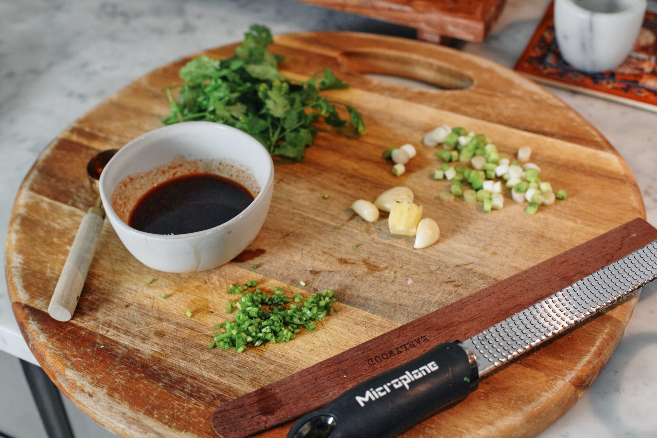a wood cutting board eith a bowl of sauce, cilantro, chopped cilantro stems, garlic gloves, a knob of ginger, chopped green onions, a wood spoon, and a microplane grater