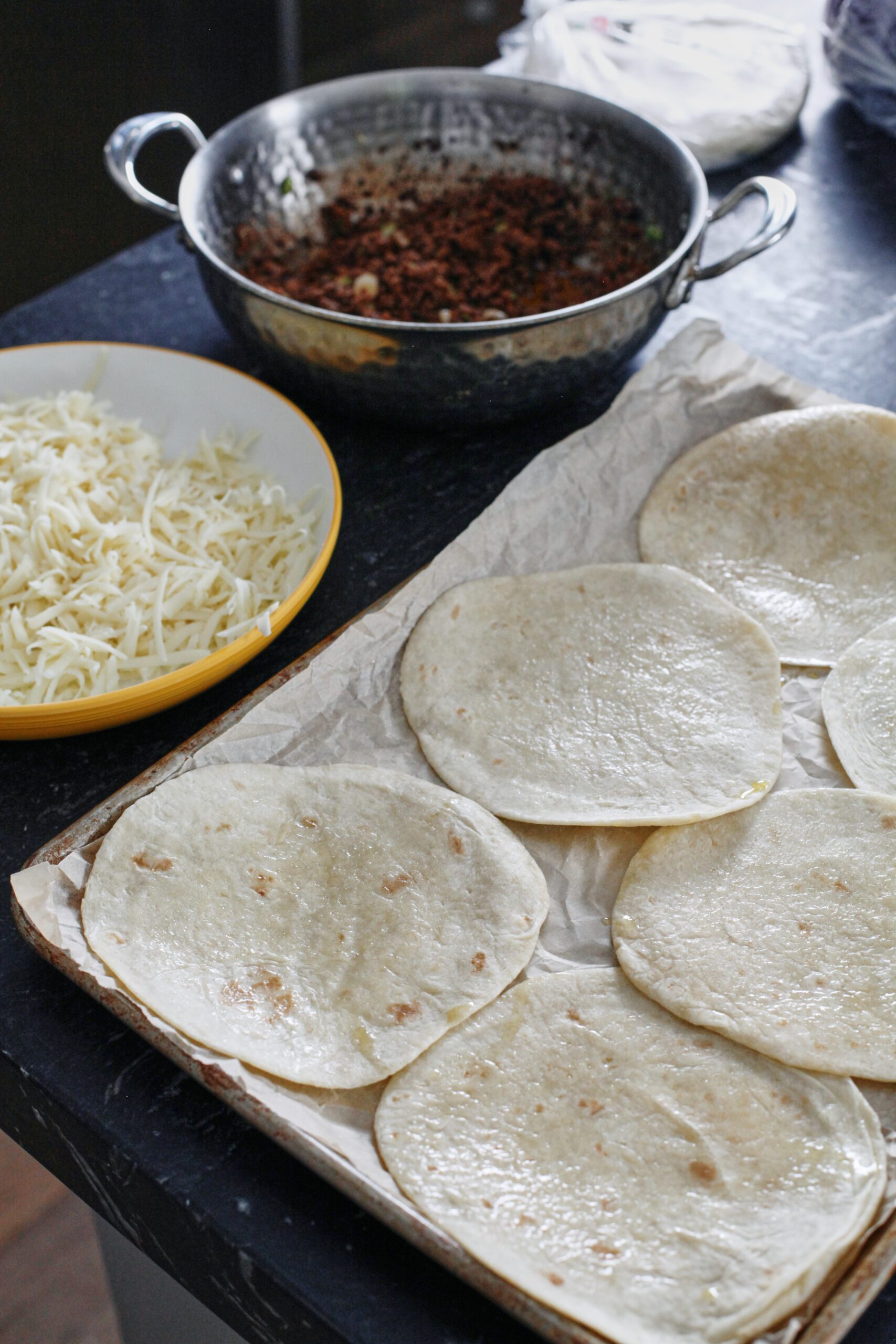 tortillas arranged on a sheet pan, a plate of shredded mozzarella, and a pan of seasoned ground beef