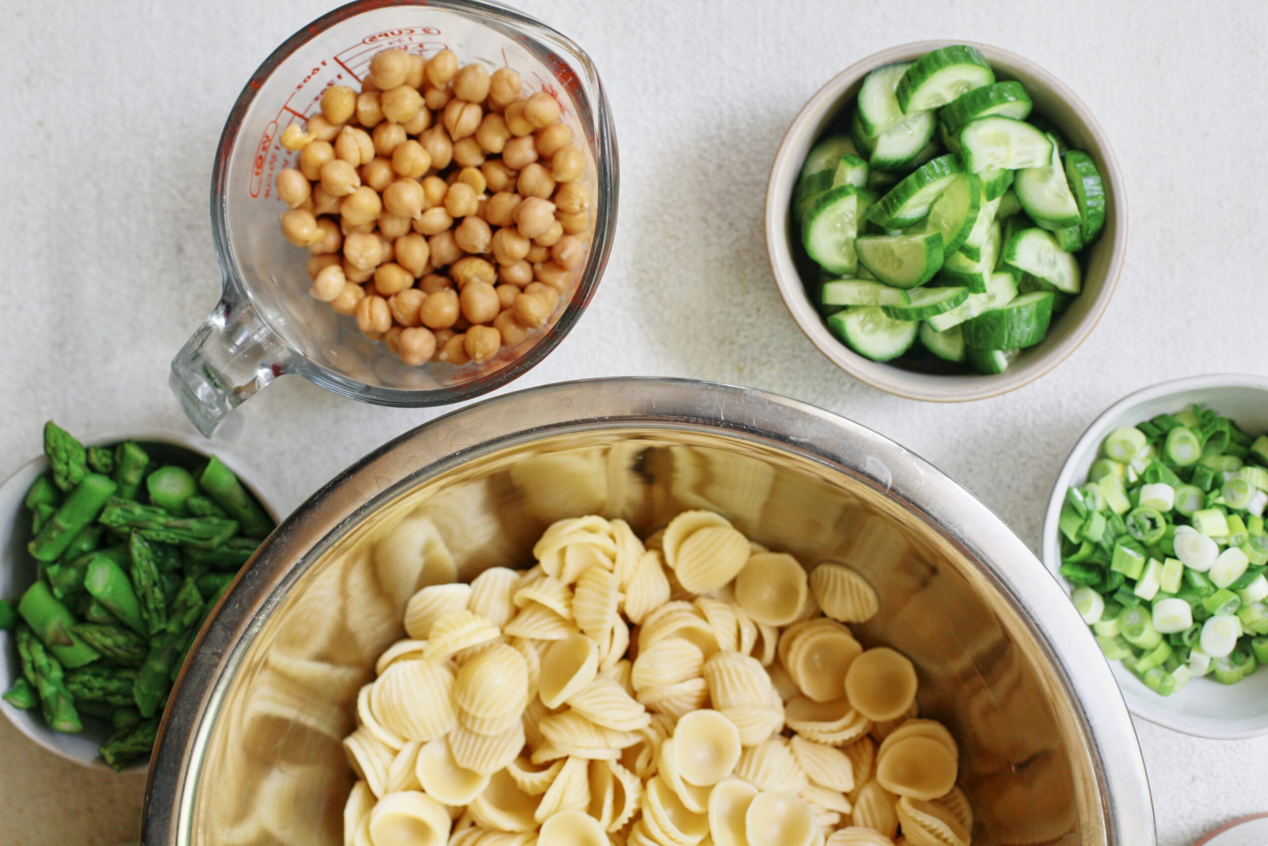 cooked orecchiette in a metal bowl surrounded by small bowls of chickpeas, cucumbers, green onions, asparagus