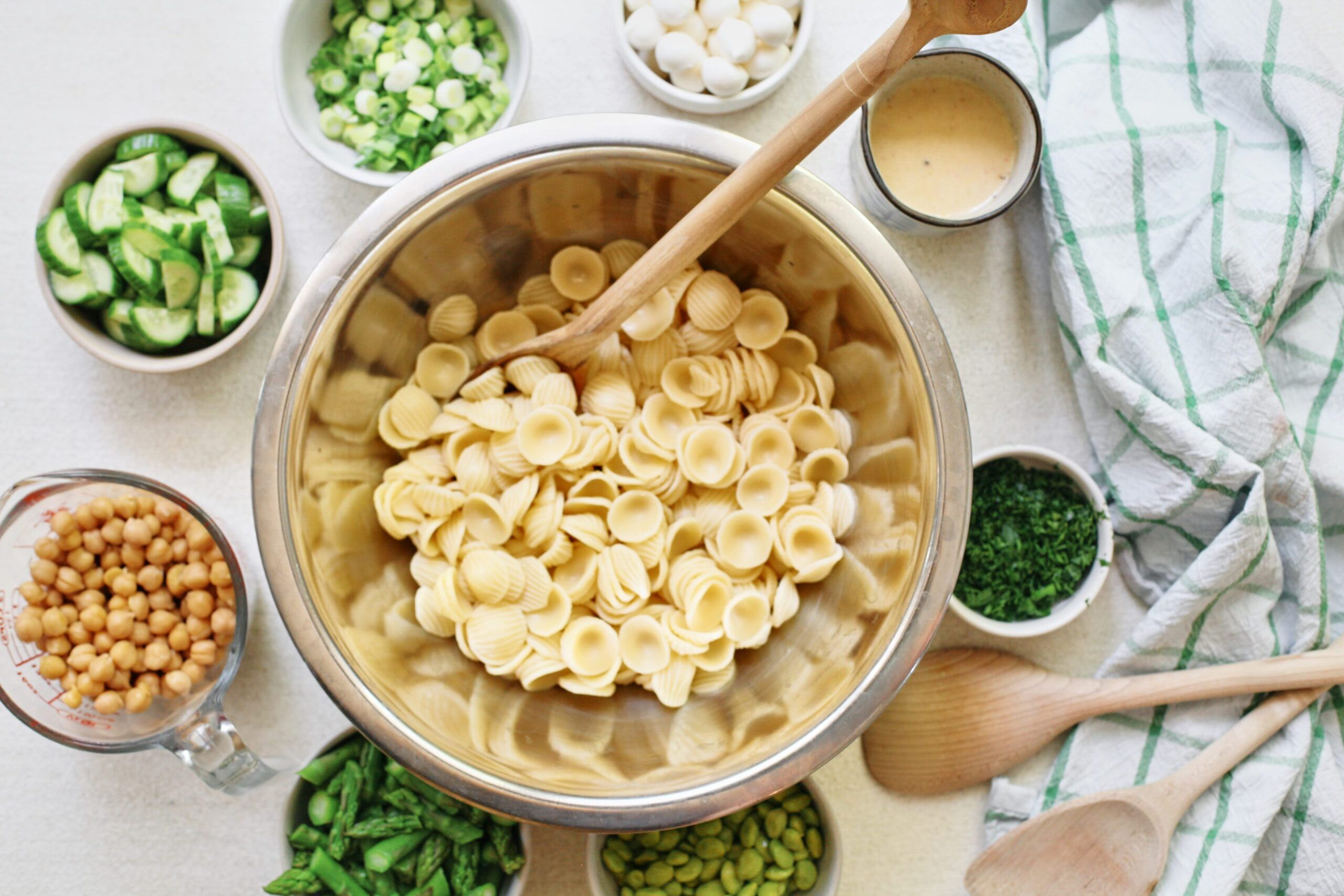 cooked orecchiette in a metal bowl surrounded by small bowls of chickpeas, cucumbers, green onions, mozzarella pearls, lemon vinaigrette, parsley, edamame, and asparagus