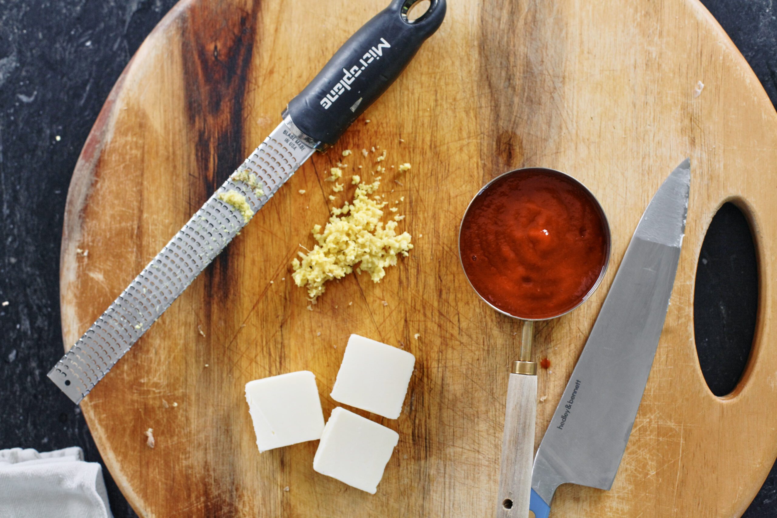 a cutting board with a grater and grated ginger, a knife and three pats of butter, and a measuring cup of tomato passata