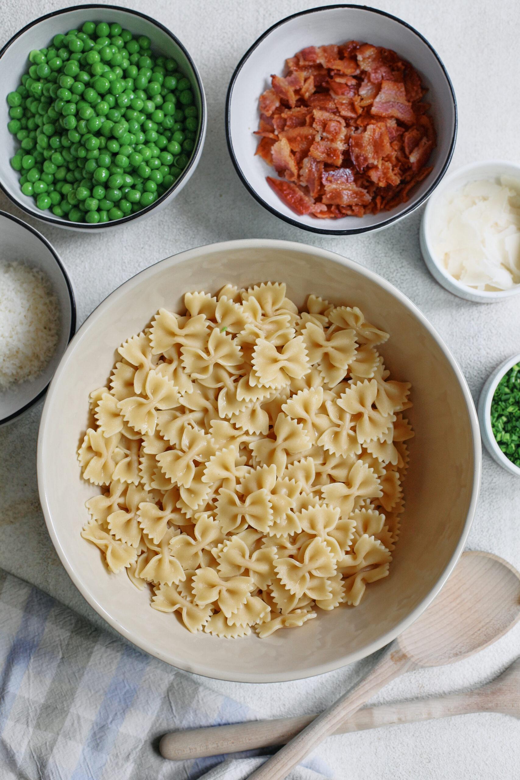 a large bowl of mini farfalle pasta surrounded by small bowls of grated parmesan cheese, peas, chopped bacon, shaved parmesan, and chopped chives