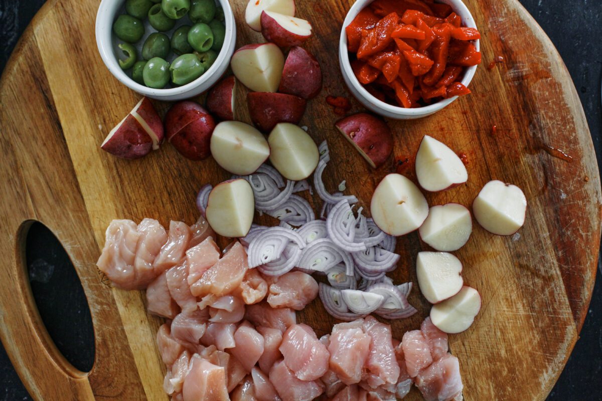 prepped ingredients on a wood cutting board: a bowl of green olives, chopped baby red potatoes, a bowl of chopped roasted red peppers, a sliced shallot, cubed chicken breast