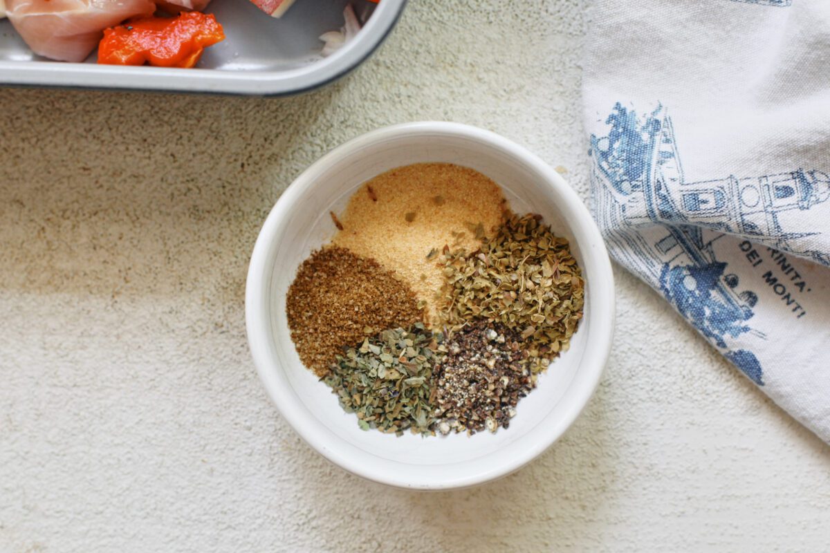dry seasonings poured into a small white bowl: garlic powder, oregano, black pepper, basil, celery salt