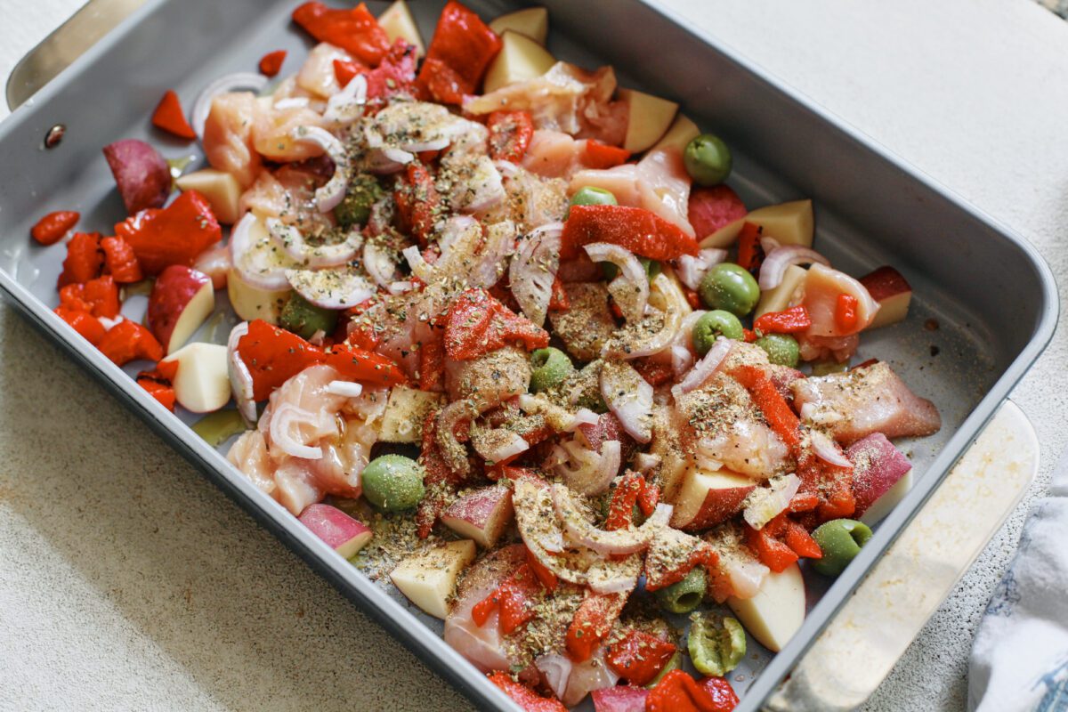 raw ingredients mixed together with seasoning blend in a baking dish: cubed chicken breast, chopped baby red potatoes, sliced shallot, green olives, chopped roasted red peppers