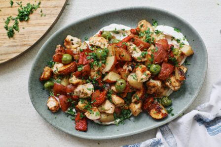 Mediterranean Chicken & Potato Tray Bake (cubed chicken breast, chopped baby red potatoes, sliced shallot, green olives, and chopped roasted red peppers) served over a yogurt sauce on an oval teal-coloured platter