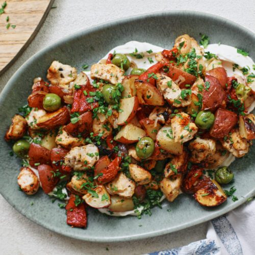 Mediterranean Chicken & Potato Tray Bake (cubed chicken breast, chopped baby red potatoes, sliced shallot, green olives, and chopped roasted red peppers) served over a yogurt sauce on an oval teal-coloured platter
