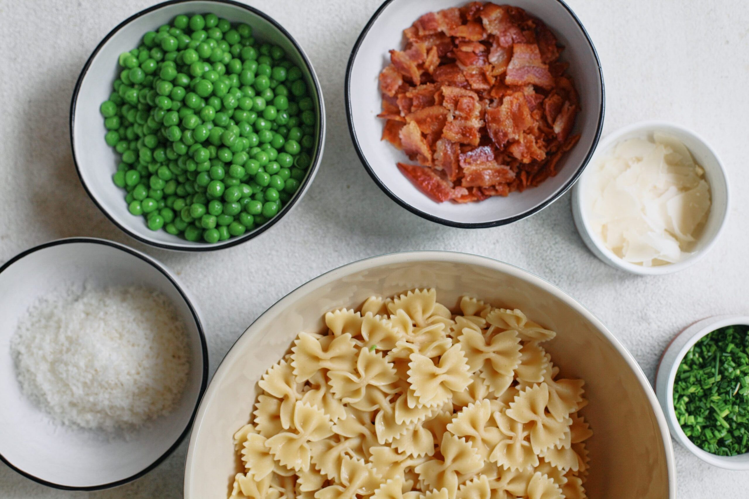 a large bowl of mini farfalle pasta surrounded by small bowls of grated parmesan cheese, peas, chopped bacon, shaved parmesan, and chopped chives