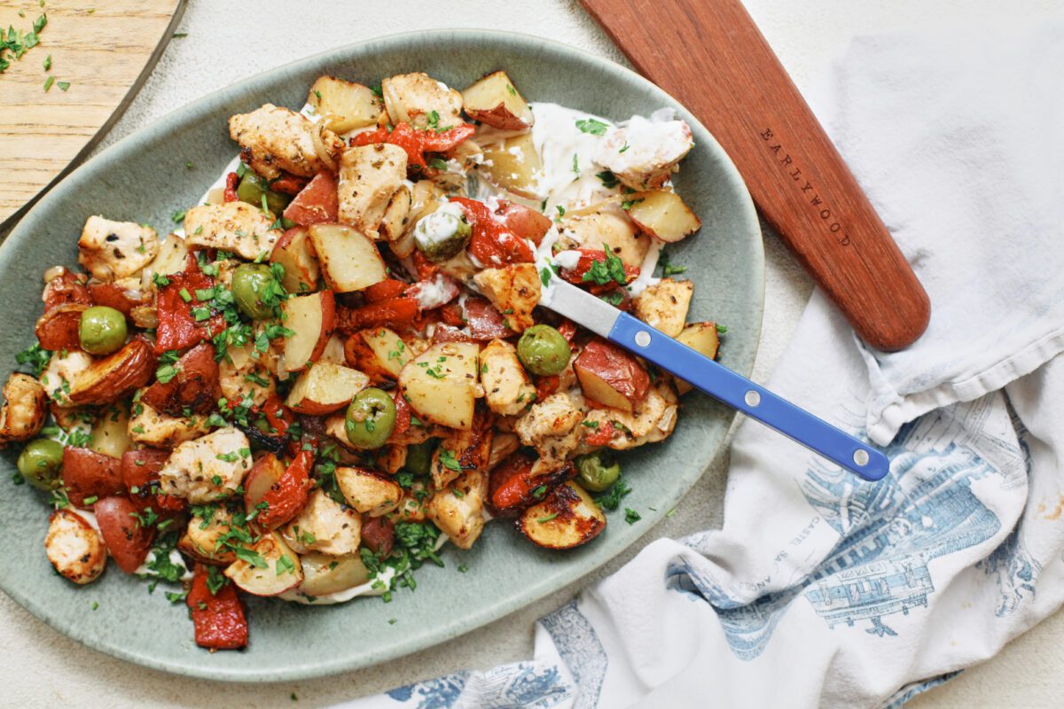 Mediterranean Chicken & Potato Tray Bake (cubed chicken breast, chopped baby red potatoes, sliced shallot, green olives, and chopped roasted red peppers) served over a yogurt sauce on an oval teal-coloured platter