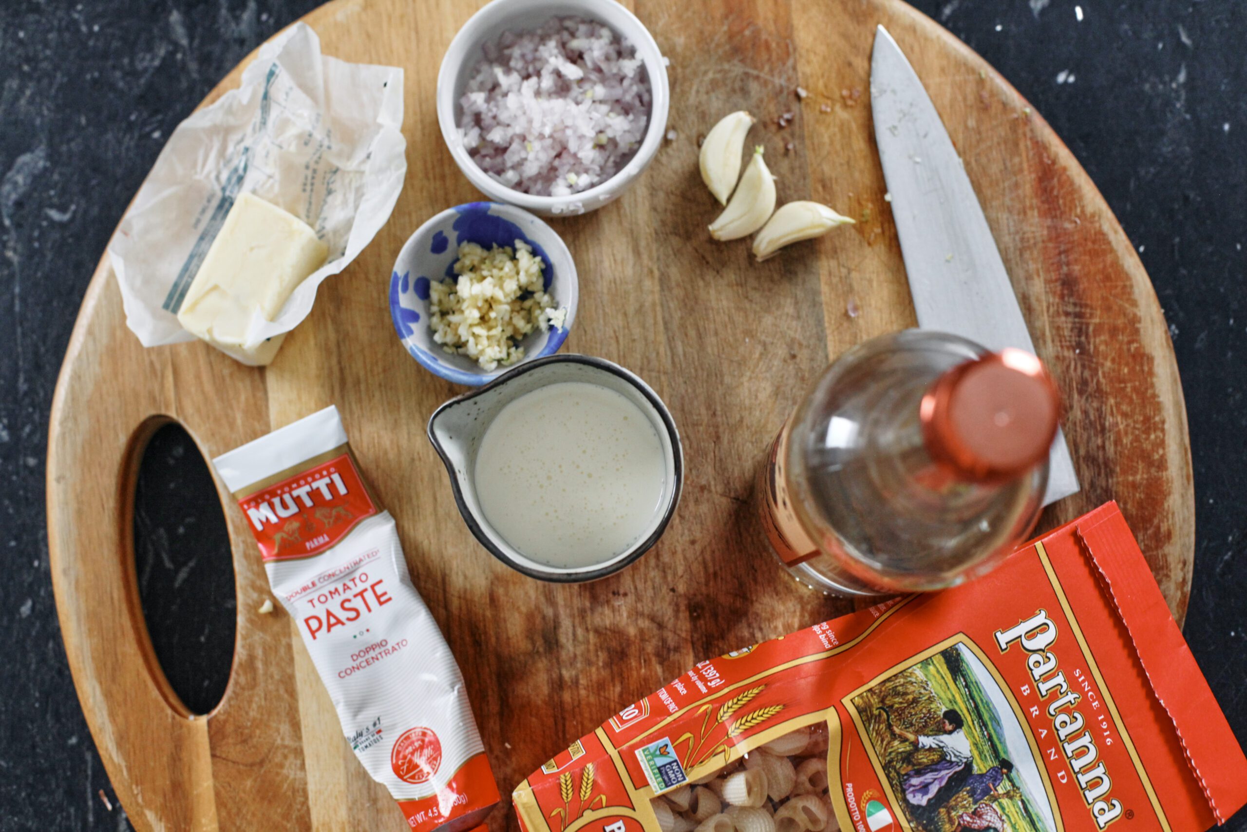 overhead photo of a round wooden cutting with ingredients for vodka pasta: half a stick of butter, a bowl of minced shallot, a bowl of minced garlic, 3 peeled garlic cloves, a tube of tomato paste, a small carafe of heavy cream, a bottle of vodka, and a bag of pipette rigate pasta