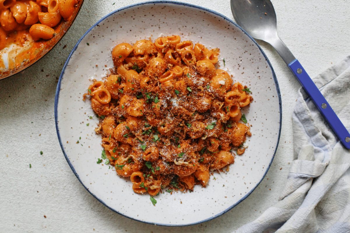 creamy vodka pasta topped with toasted garlic breadcrumbs, in a speckled white bowl with a blue-handled spoon to the side of the bowl