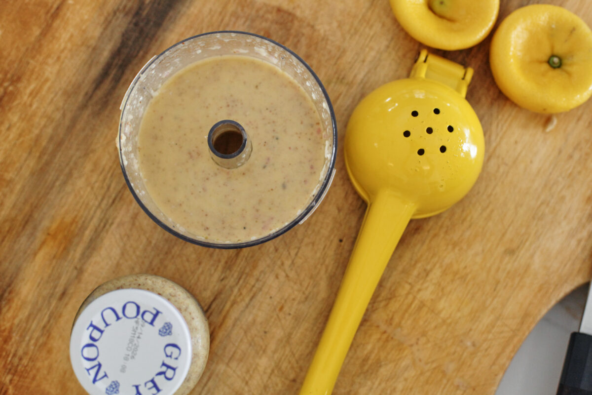 overhead photo of salad dressing in a food processor bowl, a lemon juicer and juiced lemon, and a jar of Grey Poupon mustard on a wooden cutting board