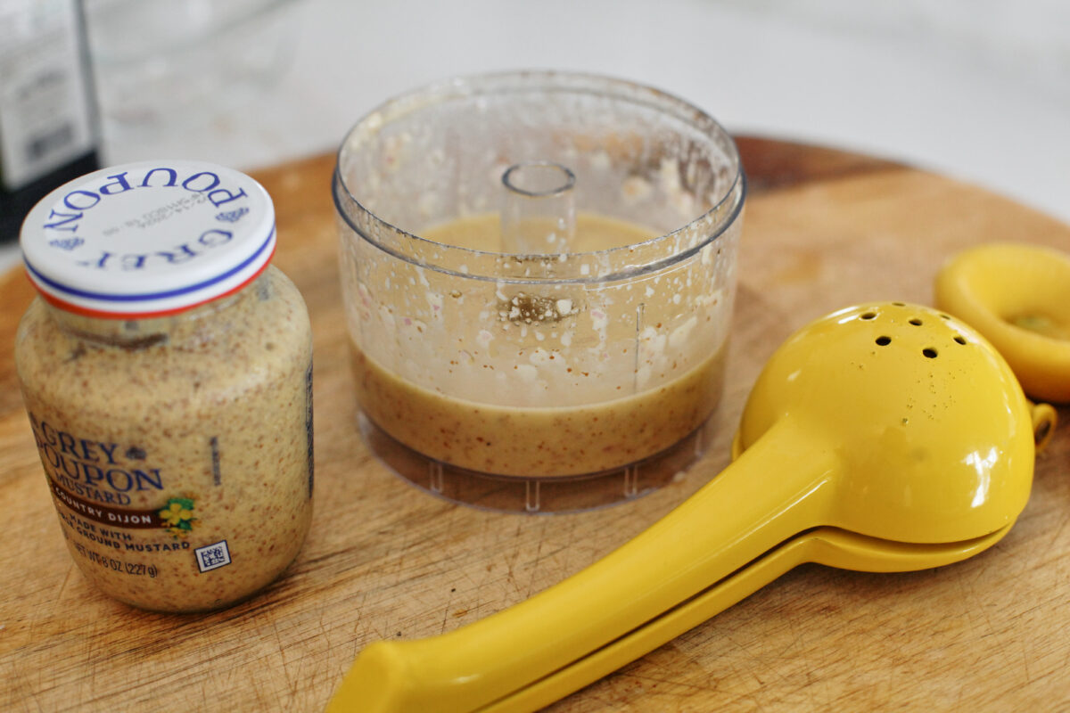 salad dressing in a food processor bowl, a lemon juicer and juiced lemon, and a jar of Grey Poupon mustard on a wooden cutting board