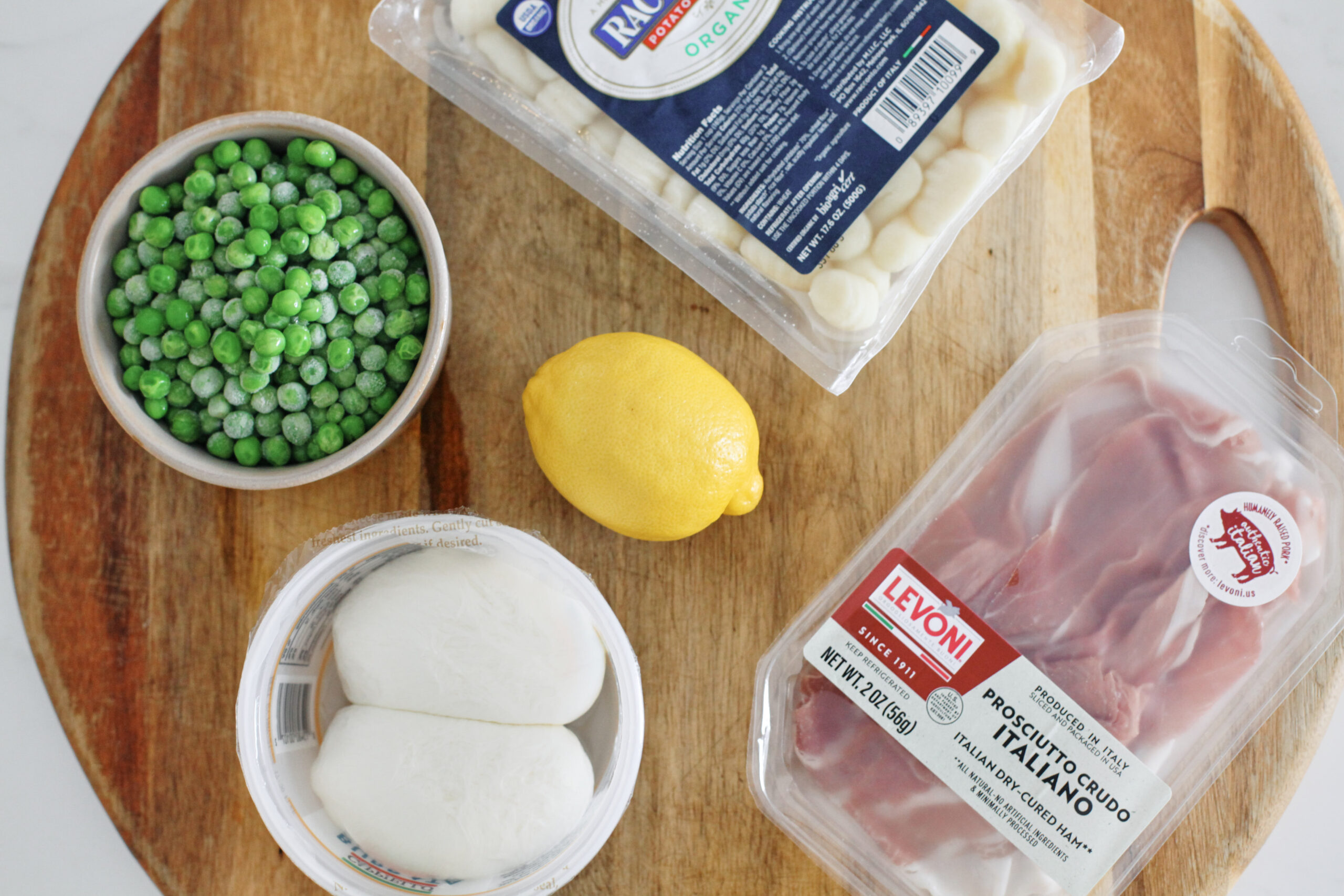 ingredients on a wooden cutting board: a bowl of frozen peas, a package of gnocchi, a lemon, two balls of burrata in a round container, and a package of prosciutto