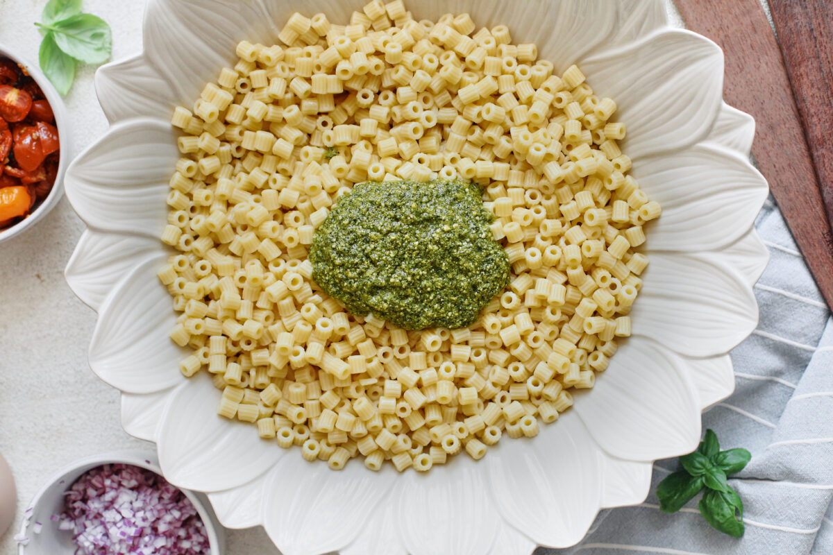 A spoonful of basil pesto added to cooked ditalini pasta in a white serving bowl