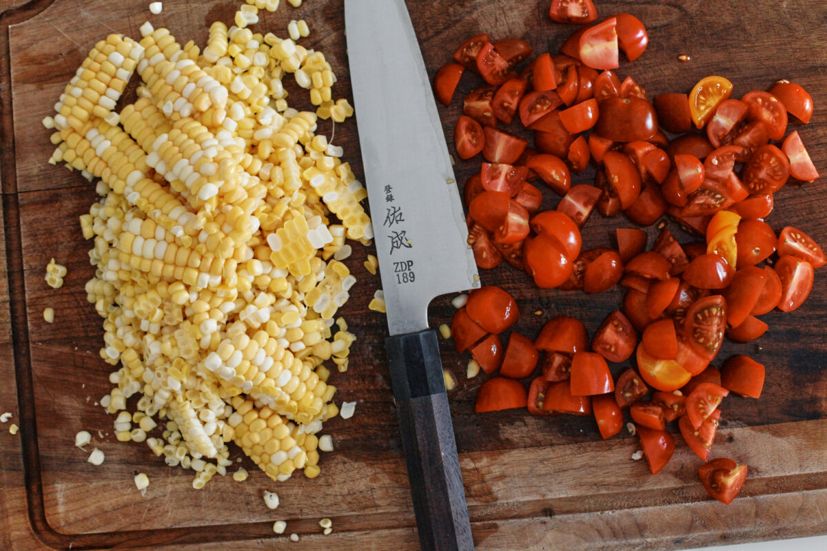 Fresh corn and chopped cherry tomatoes arranged on either side of a chef’s knife on a wooden cutting board