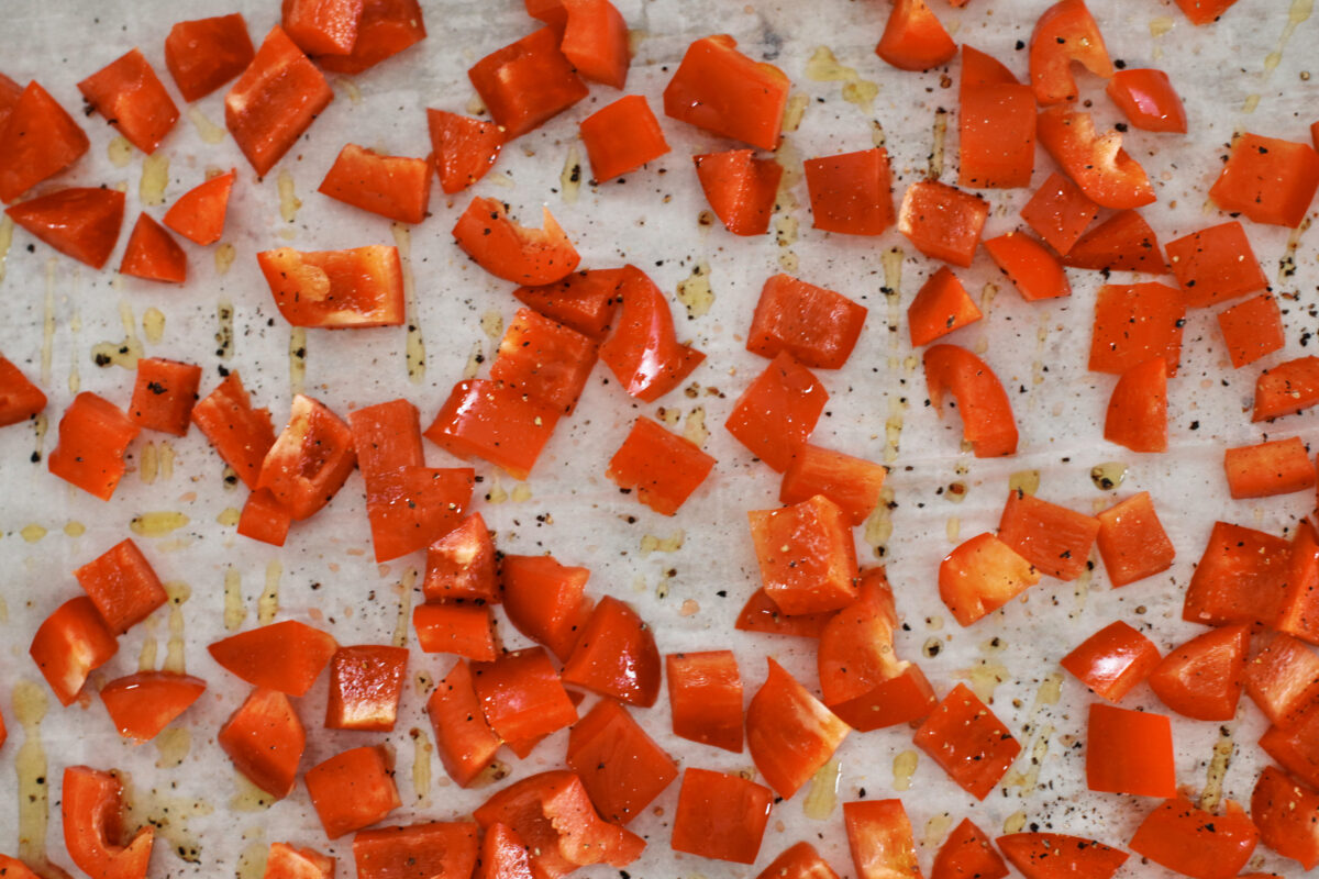 Overhead view of diced red bell peppers seasoned with olive oil, salt, and black pepper, arranged in a single layer on a parchment-lined baking sheet before baking