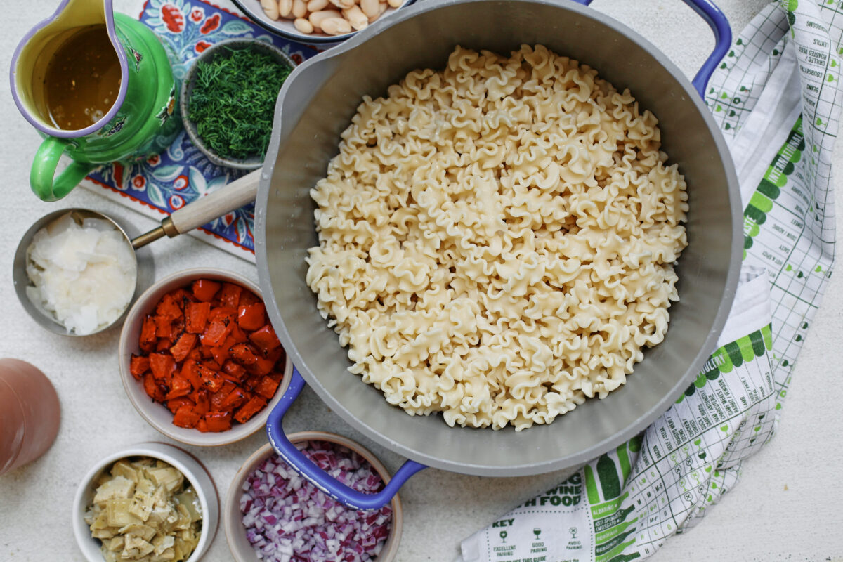 Large pot of cooked curly pasta surrounded by bowls of roasted red peppers, diced red onion, cannellini beans, chopped artichoke hearts, fresh dill, shaved Parmesan, and homemade vinaigrette