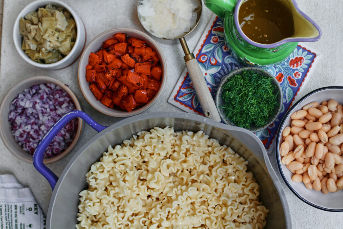 Overhead prep scene with cooked pasta and measured ingredients for antipasto pasta salad, including roasted red peppers, cannellini beans, artichoke hearts, red onion, dill, shaved Parmesan, and a small pitcher of vinaigrette
