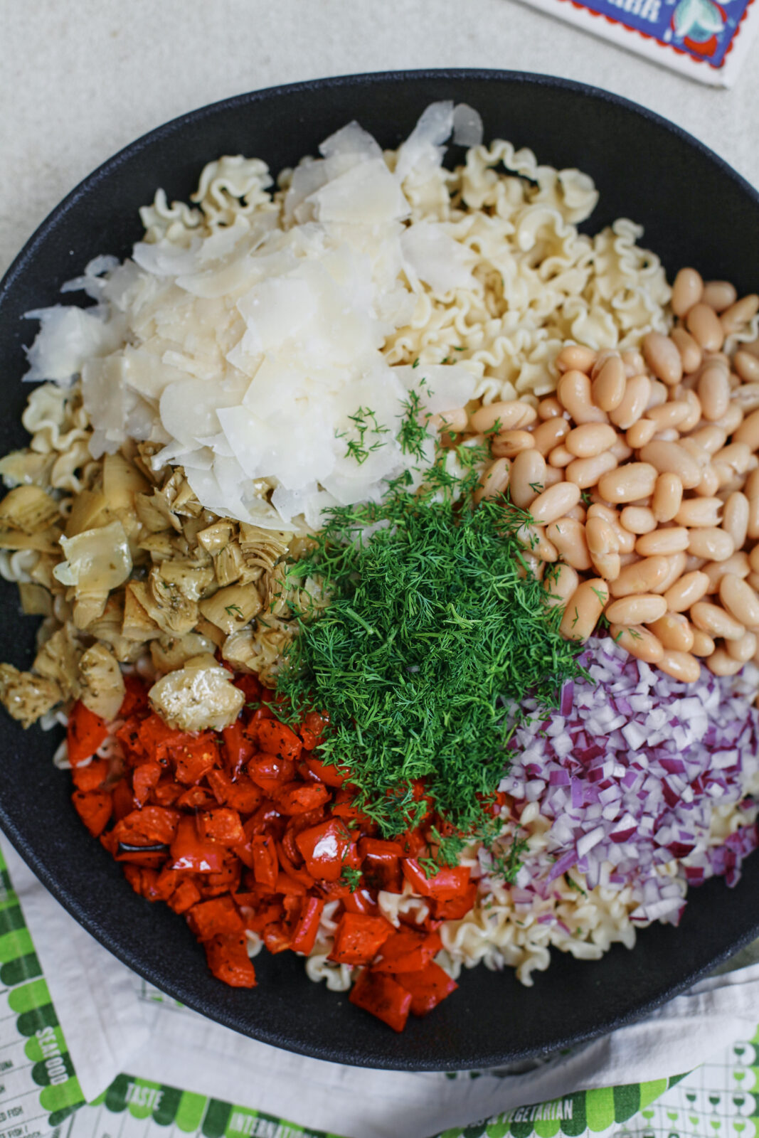 Homemade antipasto pasta salad with curly pasta, roasted red peppers, cannellini beans, marinated artichokes, red onion, fresh dill, and shaved Parmesan arranged on top before mixing
