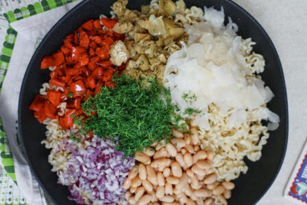 Homemade antipasto pasta salad with curly pasta, roasted red peppers, cannellini beans, marinated artichokes, red onion, fresh dill, and shaved Parmesan arranged on top before mixing
