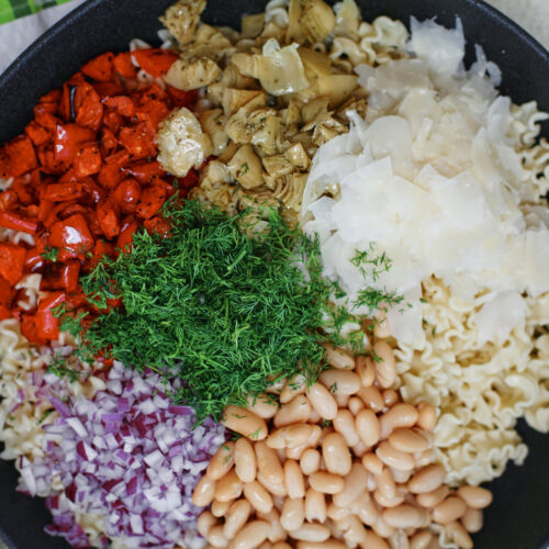 Homemade antipasto pasta salad with curly pasta, roasted red peppers, cannellini beans, marinated artichokes, red onion, fresh dill, and shaved Parmesan arranged on top before mixing