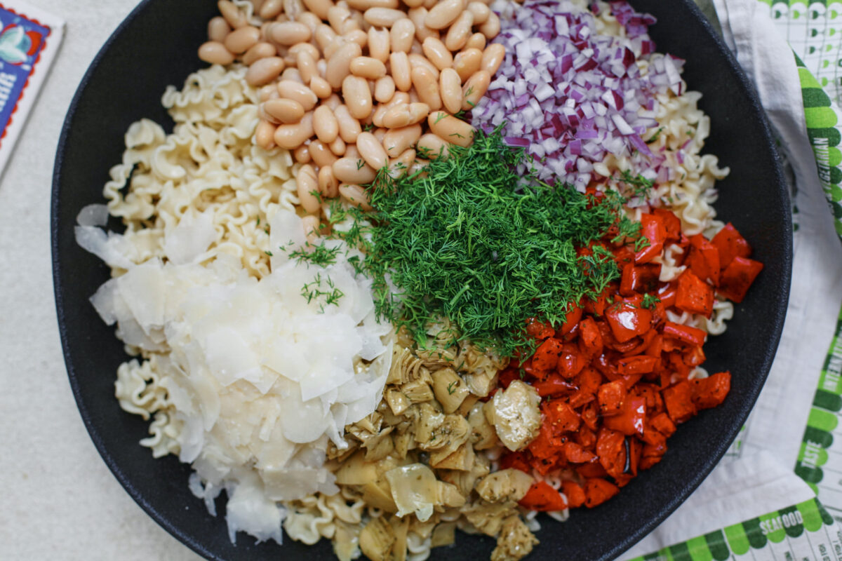 Homemade antipasto pasta salad with curly pasta, roasted red peppers, cannellini beans, marinated artichokes, red onion, fresh dill, and shaved Parmesan arranged on top before mixing