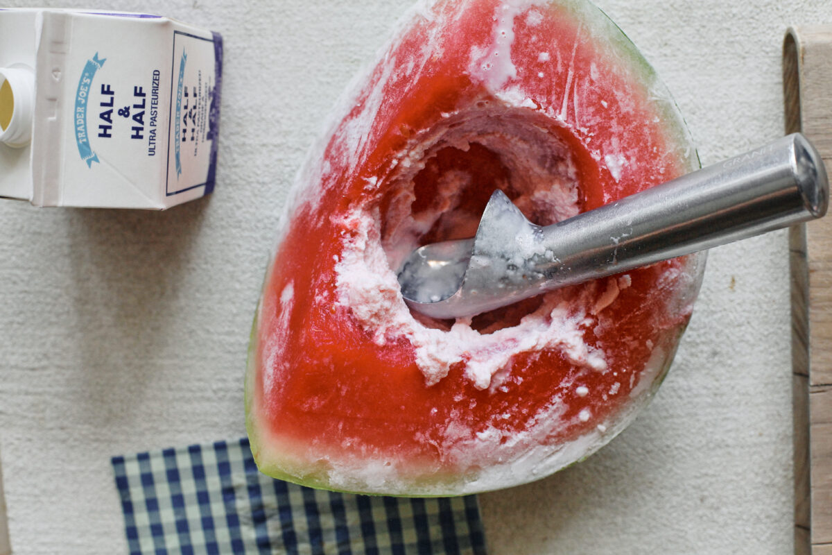 one quarter of watermelon, frozen, with cream poured into it and being scooped with an ice cream scoop