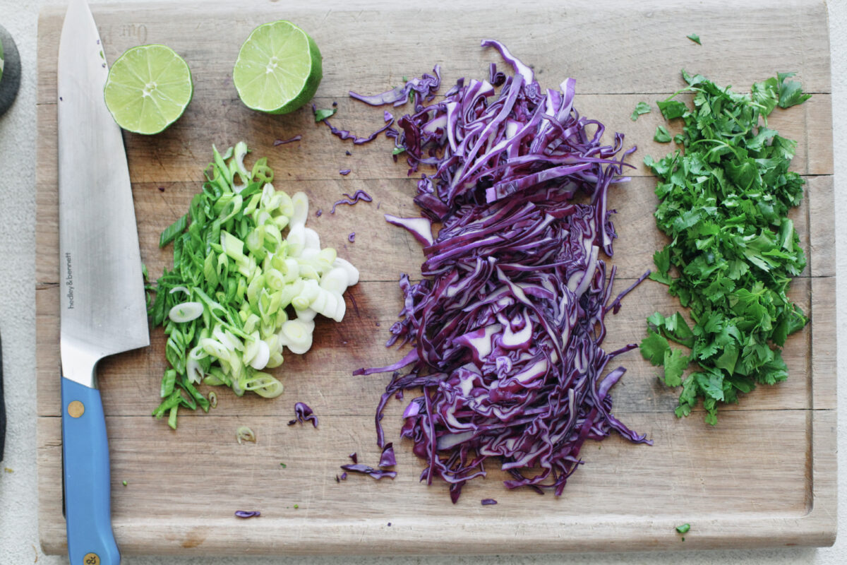 Prepped ingredients on a wood board: halved sweet potatoes, sliced purple cabbage, cilantro, green onions, and lime