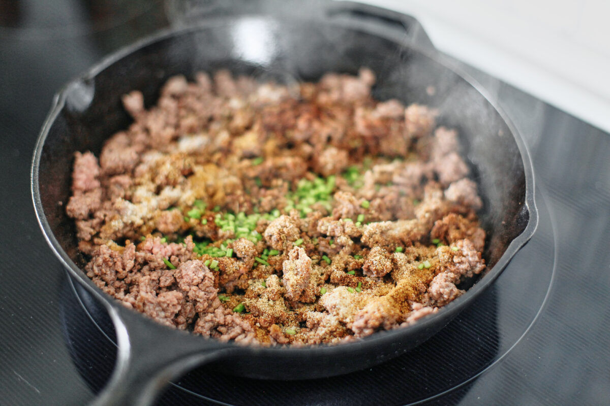 Seasoned ground beef browning in a skillet with cumin, chili powder, and chopped cilantro stems