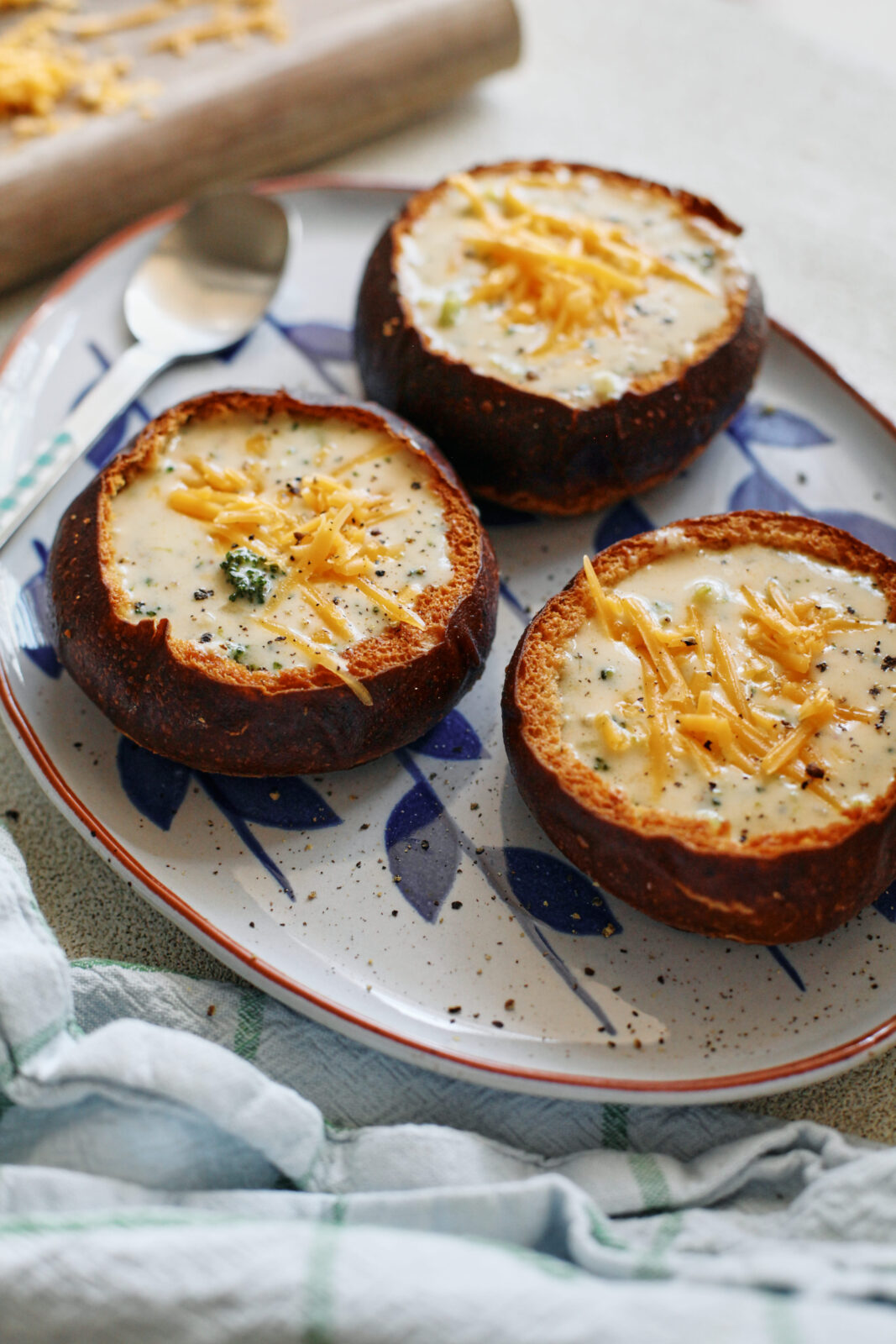 three pretzel bun bread bowls filled with broccoli cheddar soup on a white platter with blue leaf motifs