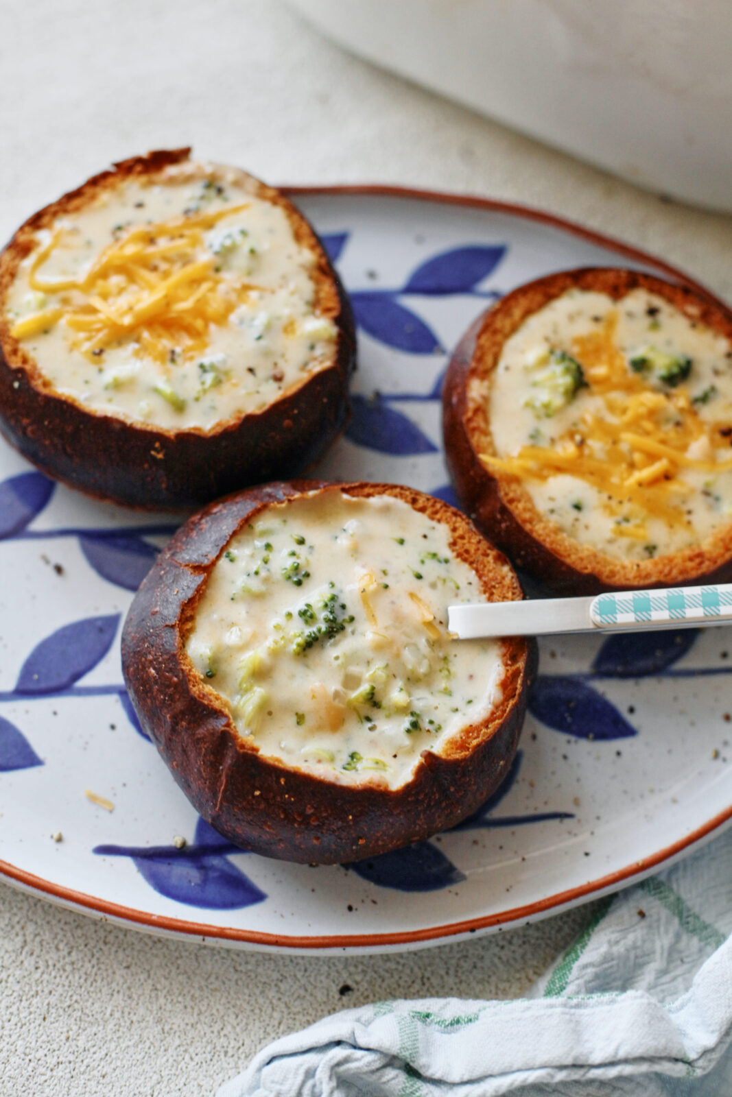 three pretzel bun bread bowls filled with broccoli cheddar soup on a white platter with blue leaf motifs
