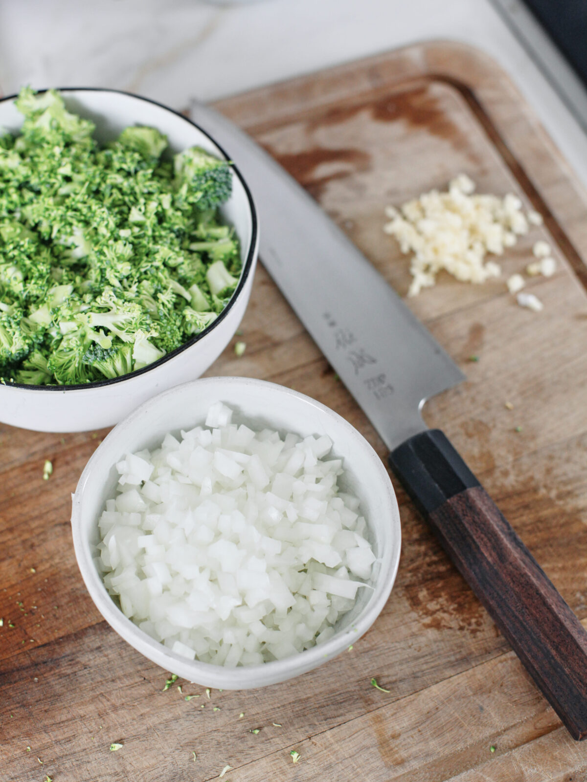 bowls of chopped onion and broccoli on a cutting board alongside a chef's knife and minced garlic