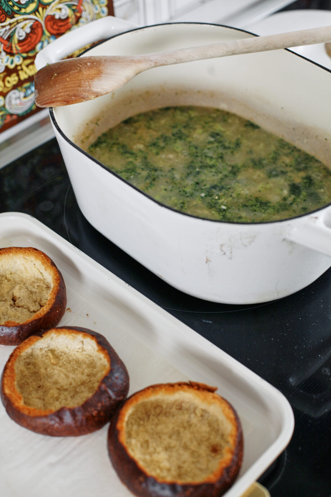 broccoli cheddar soup simmering on the stocve in a large white dutch oven next to a baking sheet with the toasted pretzel bun bowls