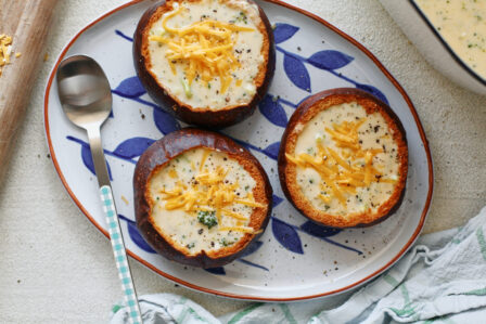 three pretzel bun bread bowls filled with broccoli cheddar soup on a white platter with blue leaf motifs