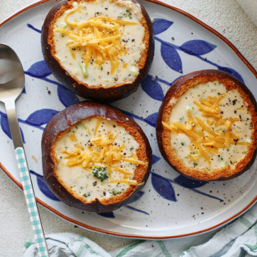 three pretzel bun bread bowls filled with broccoli cheddar soup on a white platter with blue leaf motifs