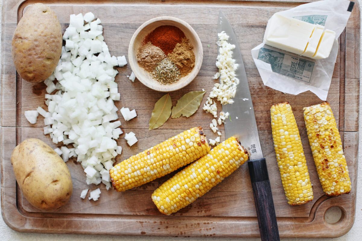 a wooden cutting board with: two potatoes, a chopped onion, a bowl of spices, two bay leaves, 4 ears of corn, a chef's knife with minced garlic on the blade, half a stick of butter
